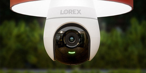 Close-up of a Lorex lightbulb security camera installed outdoors, with a wide-angle lens reflecting the surrounding area and a green indicator light glowing beneath it; blurred greenery is visible in the background.