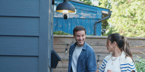 A smiling couple walks near a house entrance with a Lorex 2K Wi-Fi lightbulb security camera discreetly installed in an outdoor fixture above them.